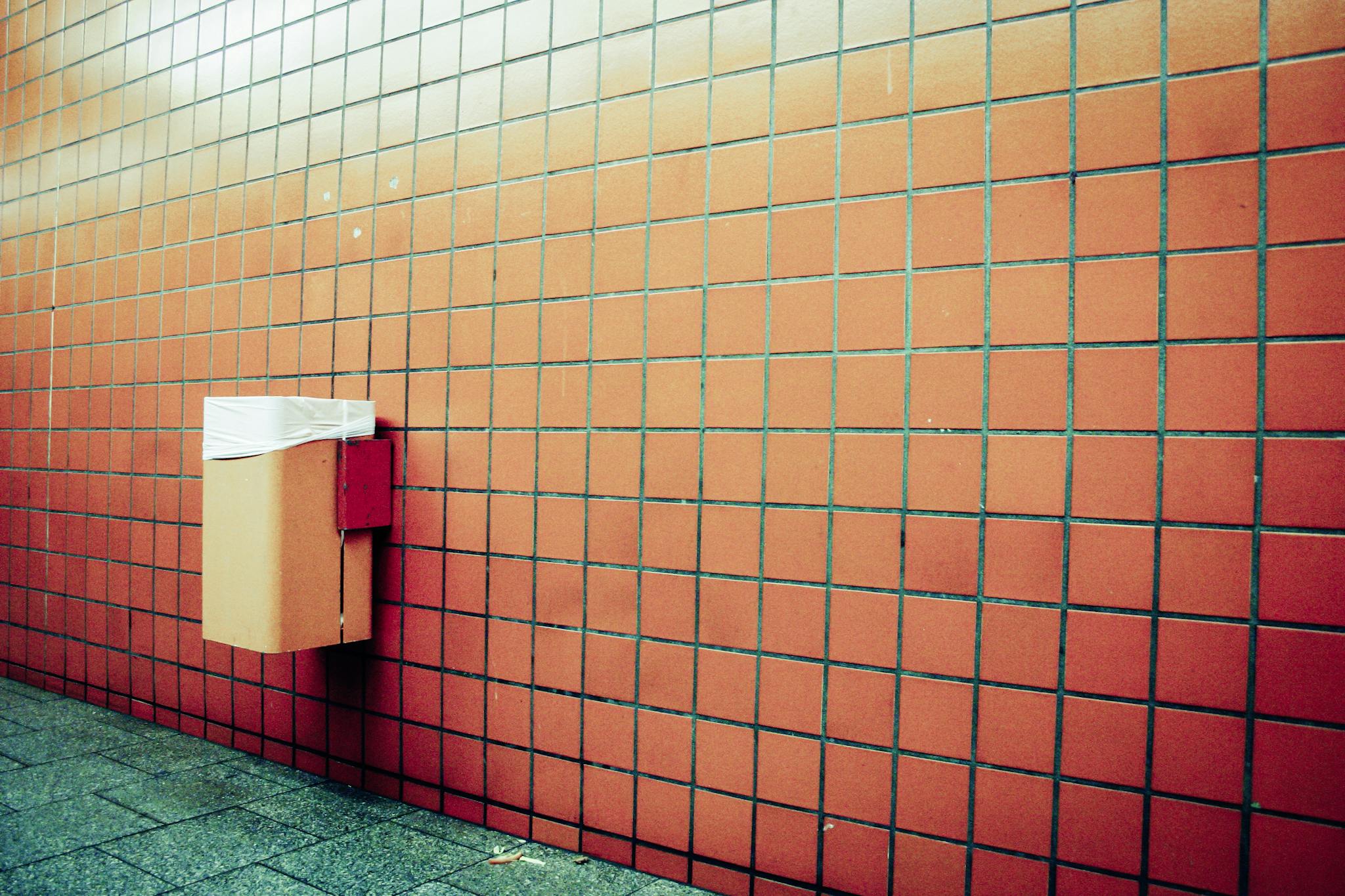 Wall-mounted garbage bin against a red tiled wall with green floor tiles.